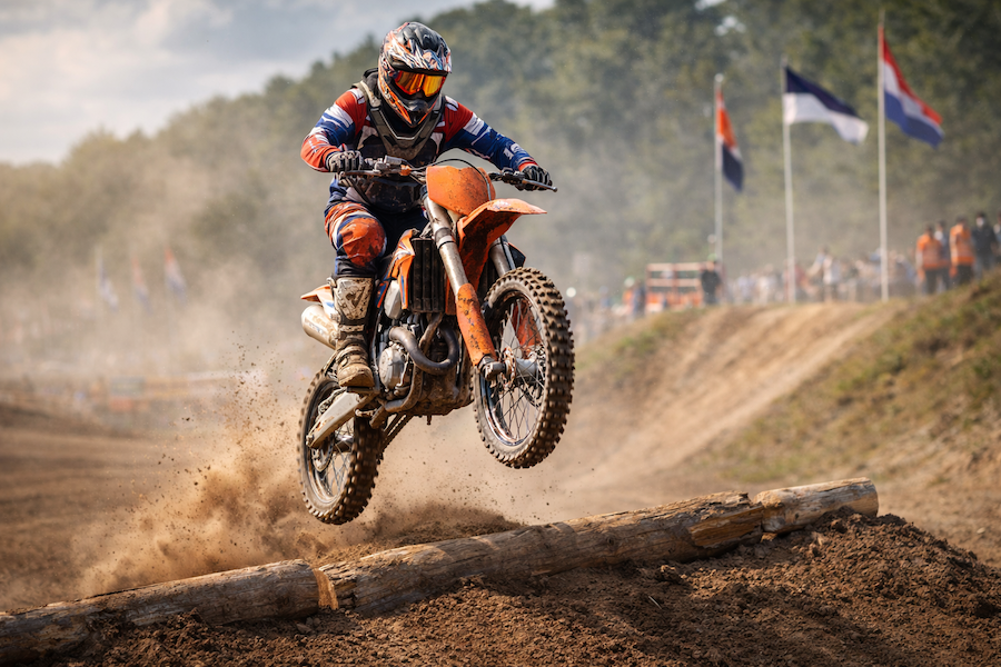 Motocrossförare hoppar över ett hinder på grusbana under tävling, med damm som virvlar bakom.